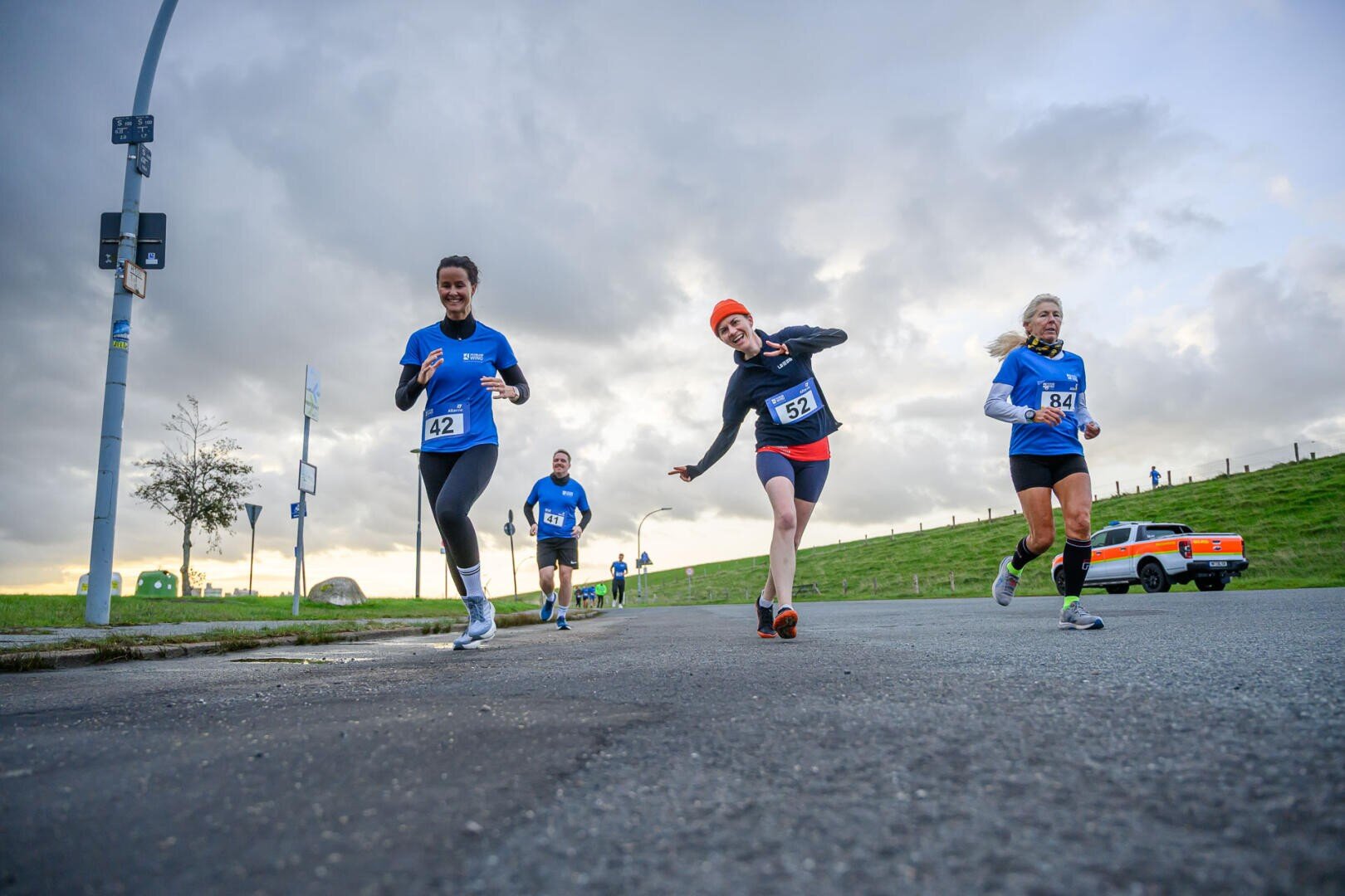 Three runners taking part in a road race, wearing blue shirts and race numbers, smiling and posing for the camera on a cloudy day. Other runners and a vintage car can be seen in the background.