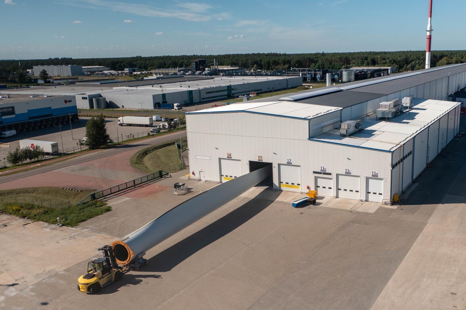 Ein großer Windturbinenflügel wird an einem klaren Tag mit einem gelben Fahrzeug aus einem weißen Industriegebäude in einer Produktionsstätte transportiert. Im Hintergrund sind Fabrikgebäude und Bäume zu sehen.
