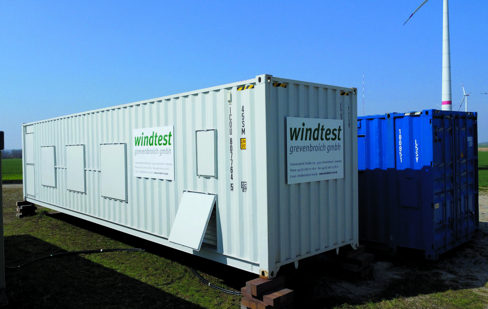 Ein großer weißer Schiffscontainer mit der Aufschrift "windtest grevenbroich gmbh" steht auf Holzklötzen neben einem blauen Container im Freien, im Hintergrund sind Windräder und offene Felder zu sehen.