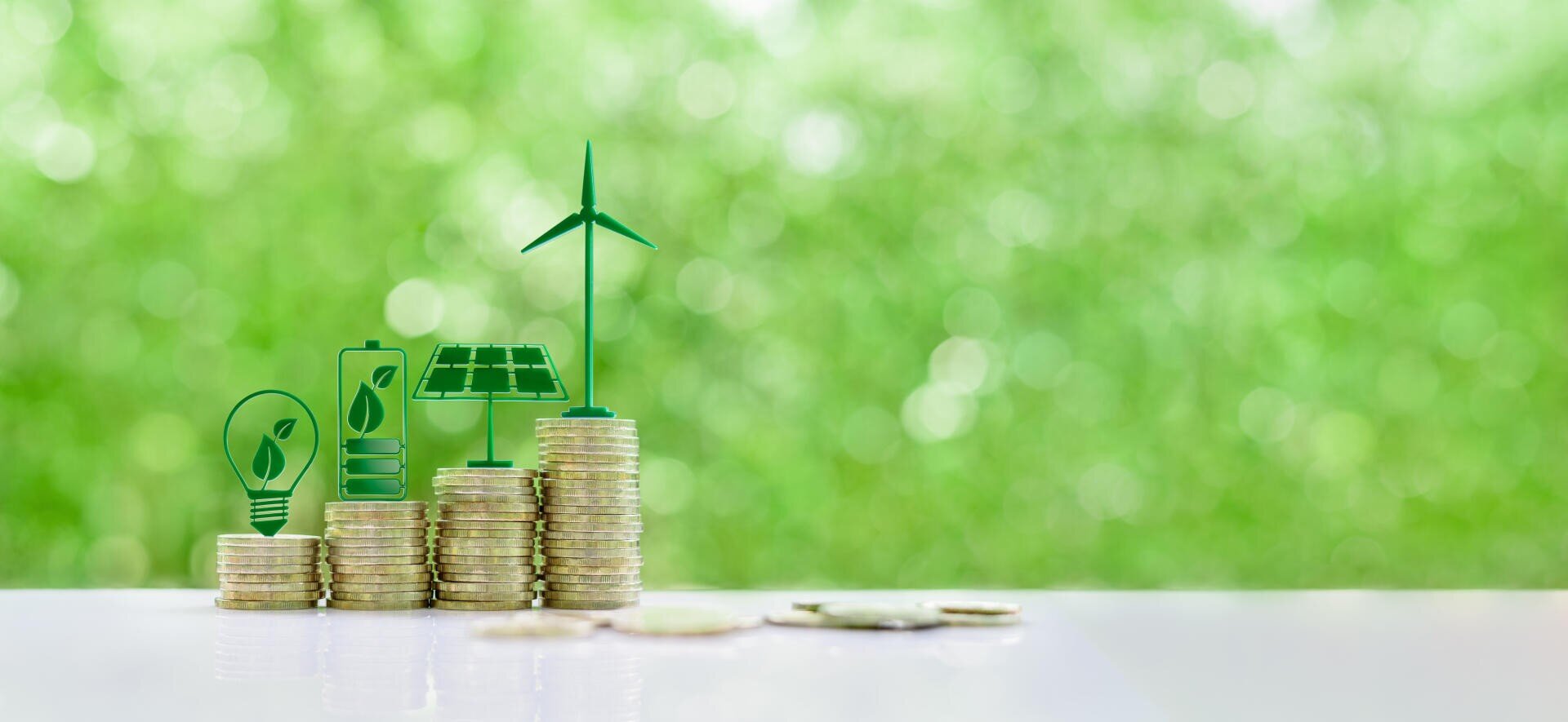 Stack of coins with green symbols representing renewable energy sources (light bulb, battery, solar panel, wind turbine) on a reflective surface, with a blurred green natural background.