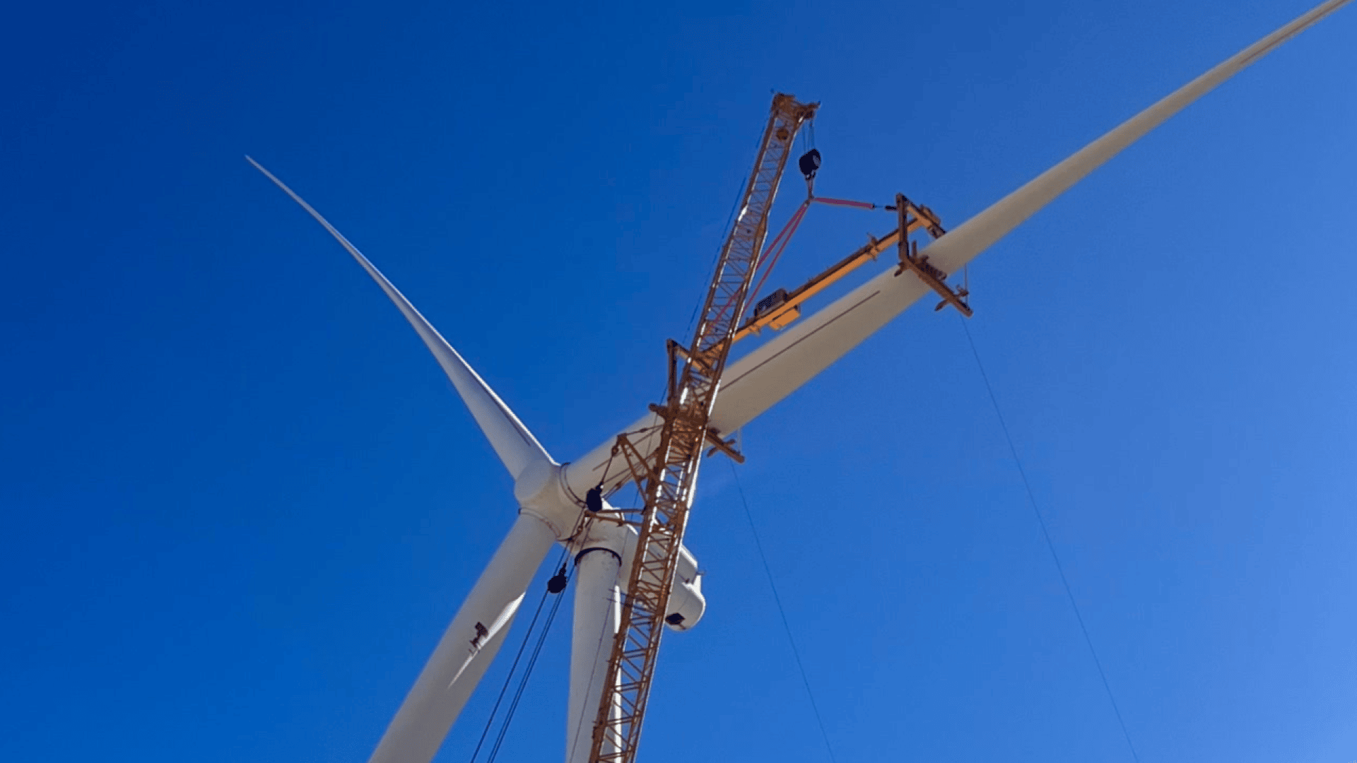 A large crane lifts a wind turbine blade for installation against a bright blue sky, while workers and equipment can be seen on the nacelle of the turbine.