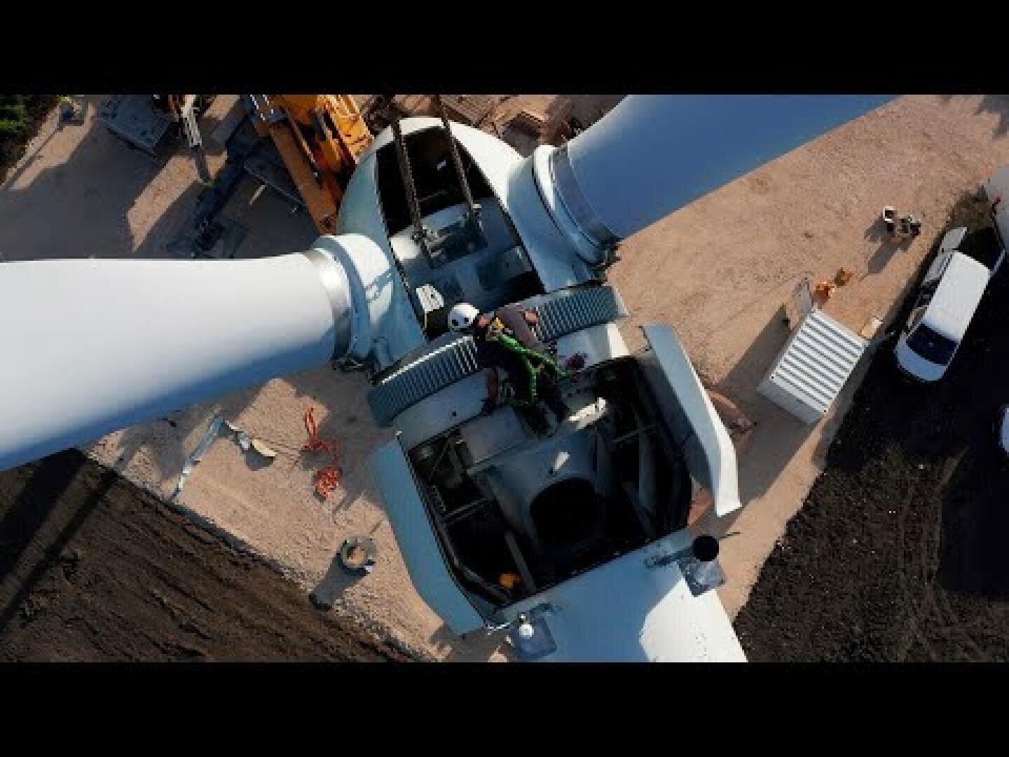 Luftaufnahme eines Arbeiters in Schutzkleidung bei Wartungsarbeiten auf einer Windkraftanlage, umgeben von großen weißen Flügeln und Baumaschinen am Boden.