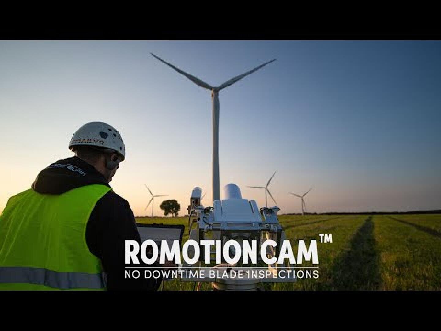 Ein Arbeiter in gelber Weste und mit Helm bedient eine Kamera oder ein Inspektionsgerät in einem Feld mit Windkraftanlagen bei Sonnenuntergang. Der Text lautet ROMOTIONCAM™ NO DOWNTIME BLADE INSPECTIONS.