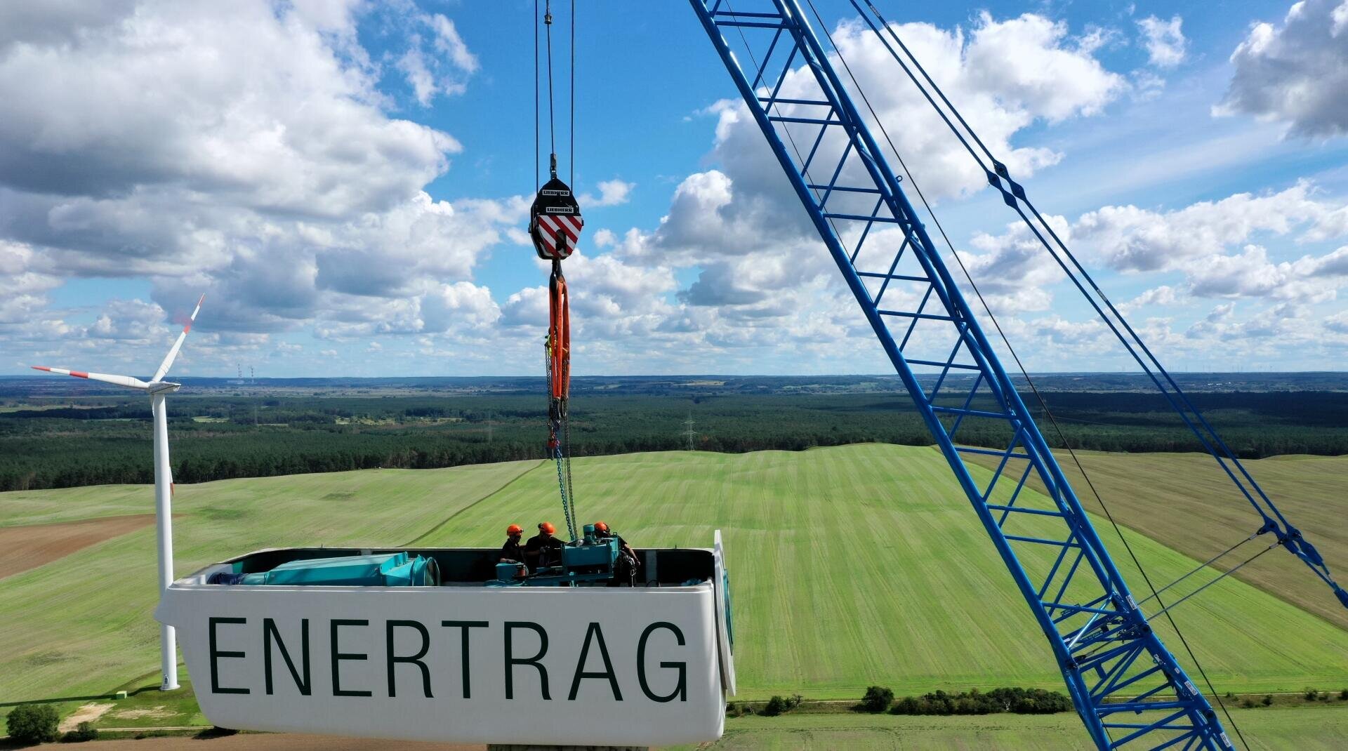 Ein großer blauer Kran hebt eine weiße Windturbinengondel mit der Aufschrift ENERTRAG über grüne Felder, in denen sich Arbeiter befinden. Im Hintergrund steht eine Windkraftanlage unter einem teilweise bewölkten Himmel.