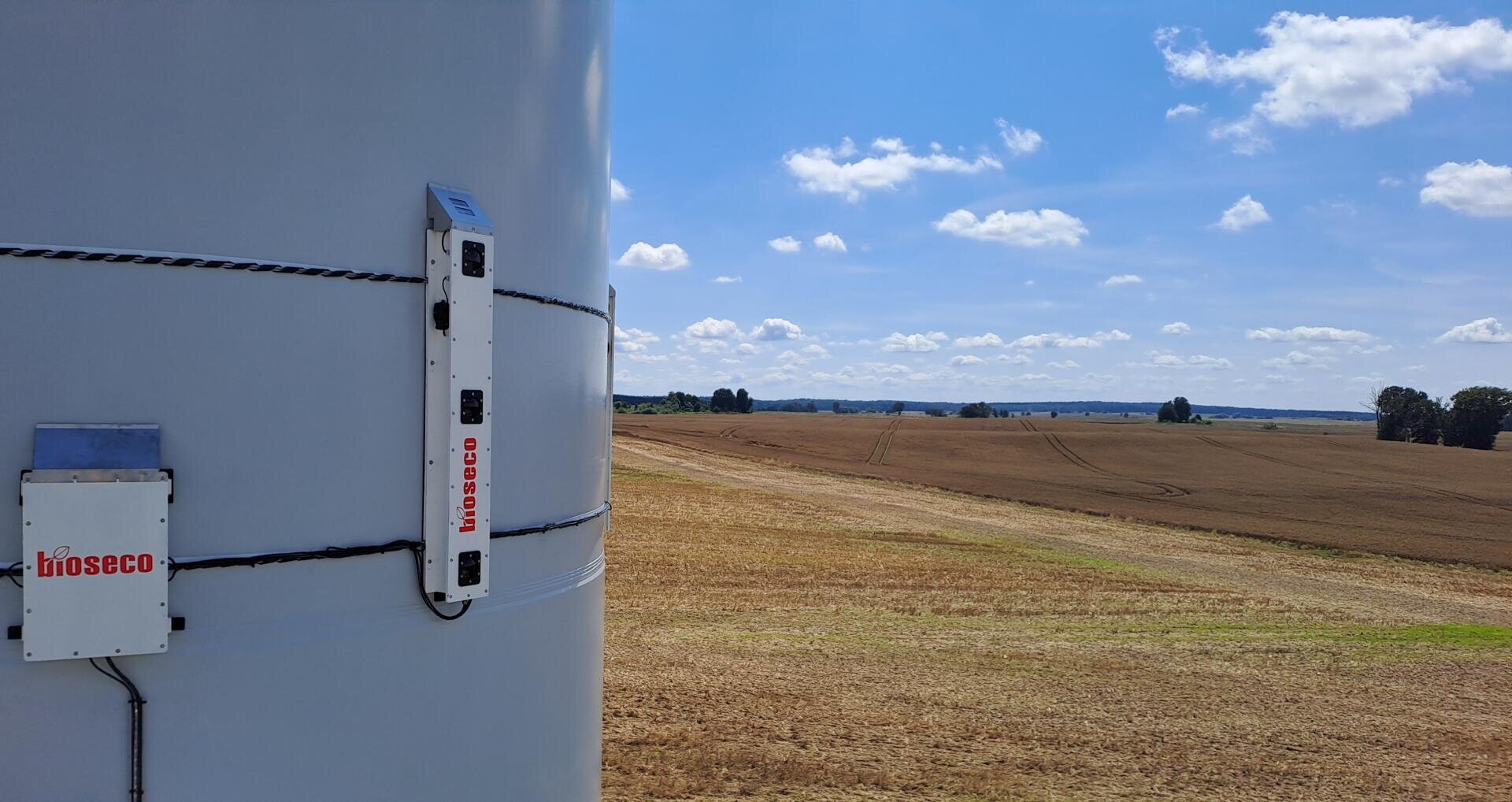 Close-up of monitoring equipment labeled bioseco mounted on a wind turbine tower overlooking a harvested field under a blue sky with scattered clouds.