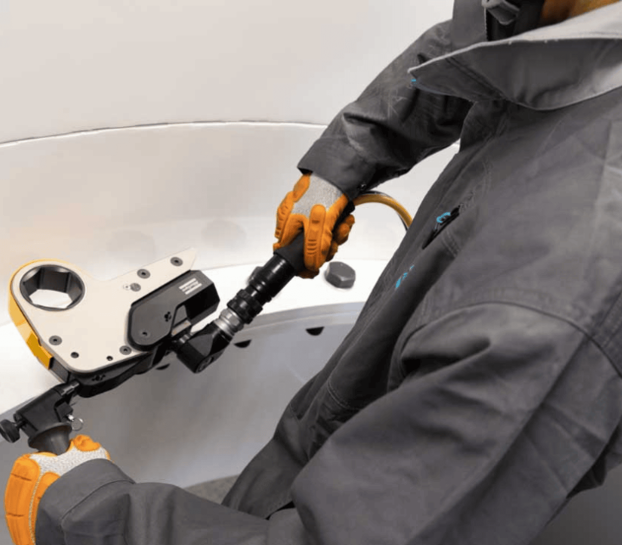 Eine Person in einem grauen Schutzanzug und orangefarbenen Handschuhen bedient einen hydraulischen Drehmomentschlüssel und zieht eine große Schraube an einer Industrieanlage fest.