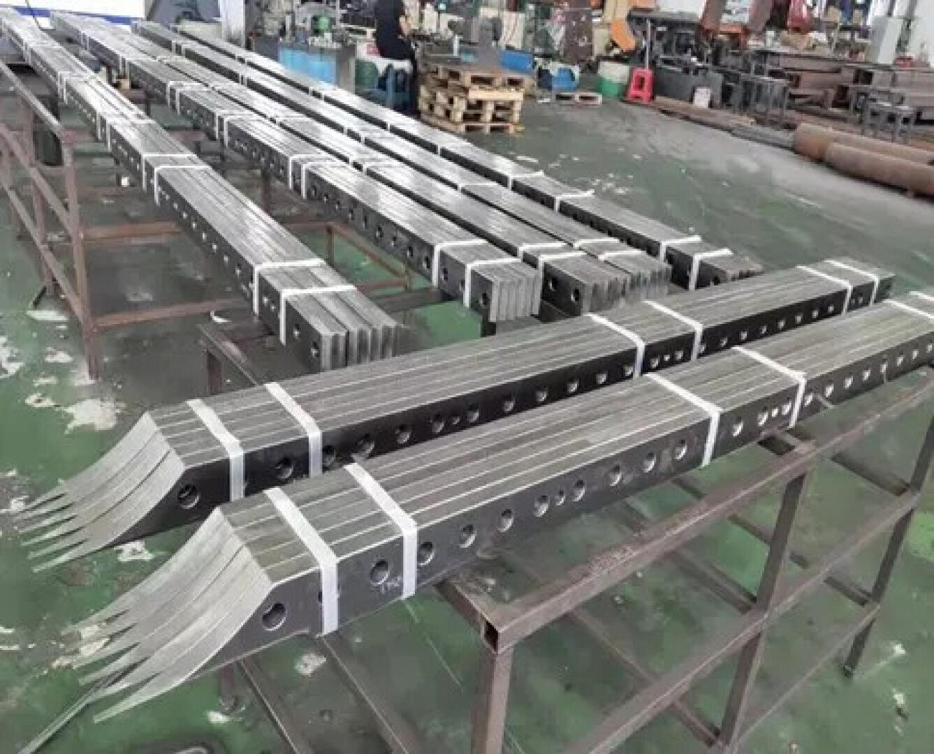 Mehrere Bündel langer, rechteckiger Metallstangen mit mehreren Löchern und gegabelten Enden sind auf Metallgestellen in einer Industriehalle gestapelt. Der Boden ist grün und im Hintergrund sind verschiedene Werkzeuge und Geräte zu sehen.