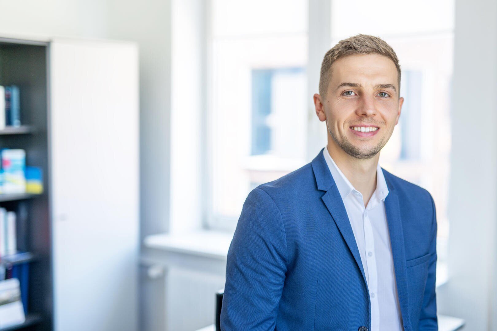 Ein Mann in blauem Blazer und weißem Hemd lächelt, während er in einem hellen Büro mit großen Fenstern und einem Bücherregal im Hintergrund steht.