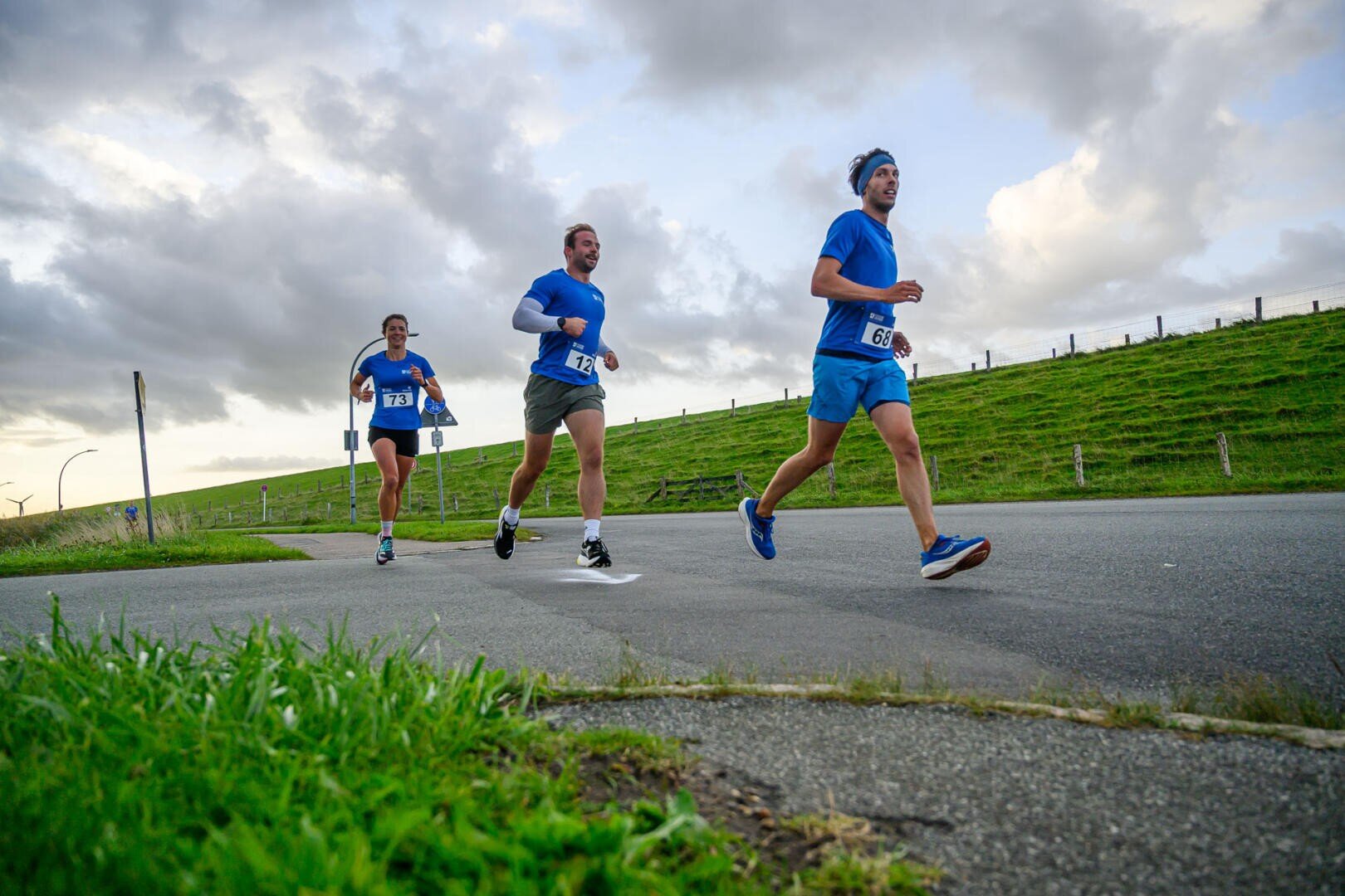 Three runners wearing blue shirts and race numbers jog along a paved road near a grassy hill under a cloudy sky, taking part in an open-air race or marathon.