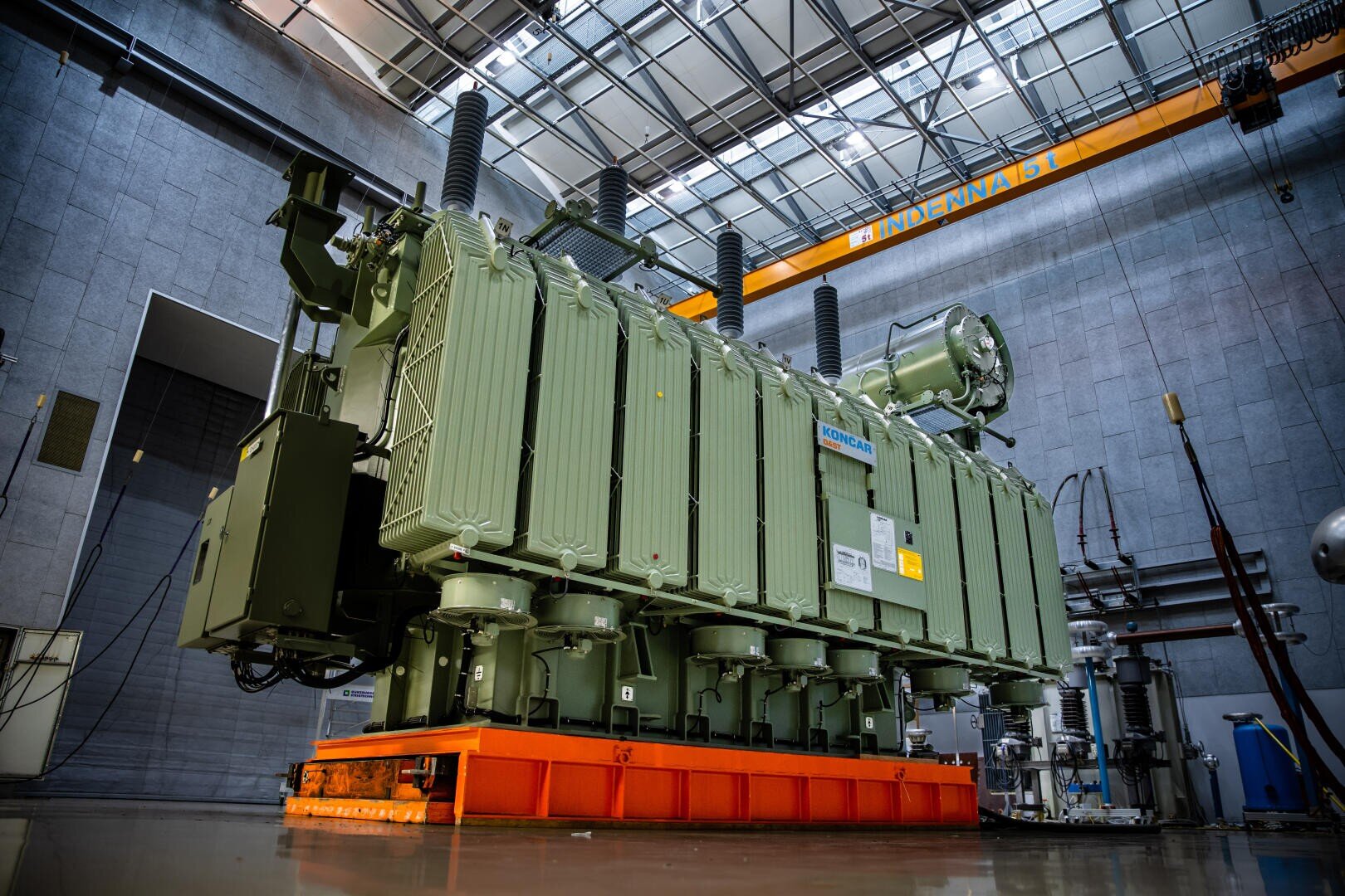 Ein großer Industrietransformator mit grünen Rippenplatten steht auf einer orangefarbenen Plattform in einem modernen Gebäude mit hohen Decken, grauen Wänden und einem gelben Brückenkran mit der Aufschrift INDARMA SL.