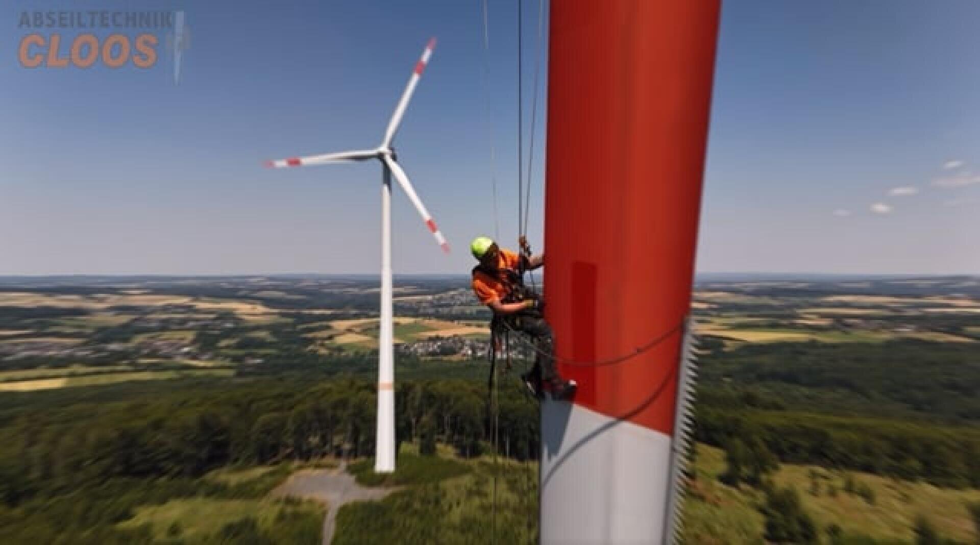 A worker in safety gear is suspended by ropes, inspecting or maintaining a large red and white wind turbine. Another wind turbine stands in the background, with fields and forests visible below under a clear sky.