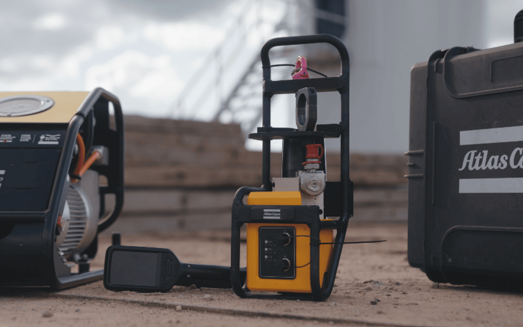 A yellow industrial device with dials and controls is set on a sandy surface outdoors, next to black equipment cases. A pink toy is attached to the handle. The background shows blurred wooden planks and building structures.