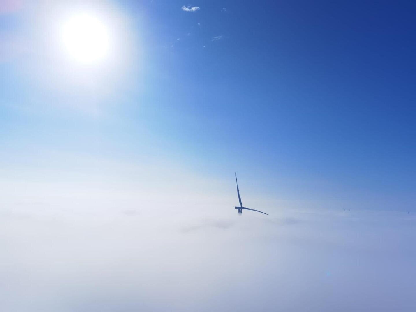 Ein einzelnes Windrad taucht durch dichte weiße Wolken hindurch auf, während über ihm die helle Sonne in einen strahlend blauen Himmel scheint. Die Szene ist friedlich und stimmungsvoll, wobei die Turbine teilweise von Nebel umhüllt ist.