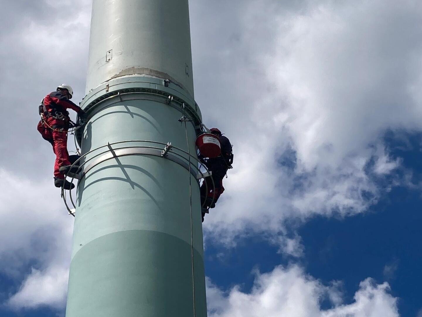Zwei Arbeiter in roter Schutzkleidung und mit Helmen klettern bei teilweise bewölktem Himmel auf ein hohes, zylindrisches Bauwerk, wobei sie sich mit Gurten und Sicherungsseilen abstützen.