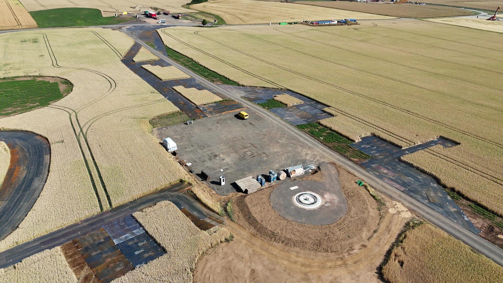 Luftaufnahme einer Baustelle in einem ländlichen Gebiet mit Geräten, Fahrzeugen und Materialien auf einer gerodeten Fläche, die von Ackerland und mobilen Baustraßen aus Stahlplatten umgeben ist.