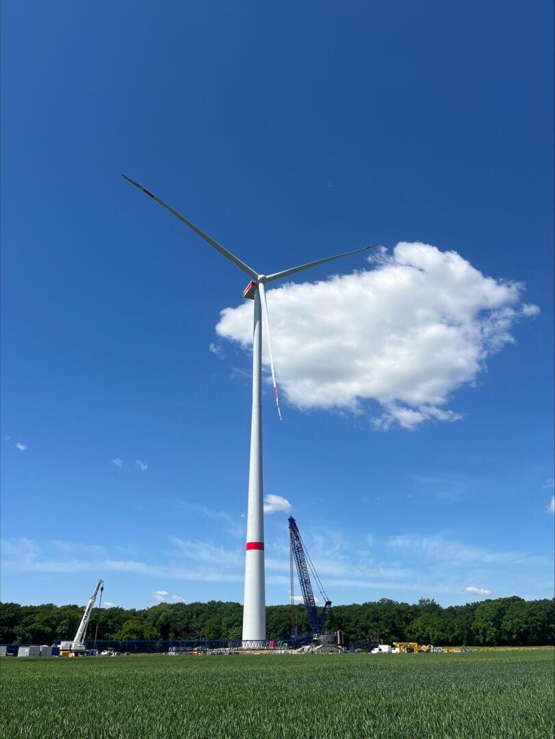 A tall wind turbine stands in a grassy field under a blue sky with a single white cloud. Construction cranes and equipment are visible near the base, with a line of trees in the background.