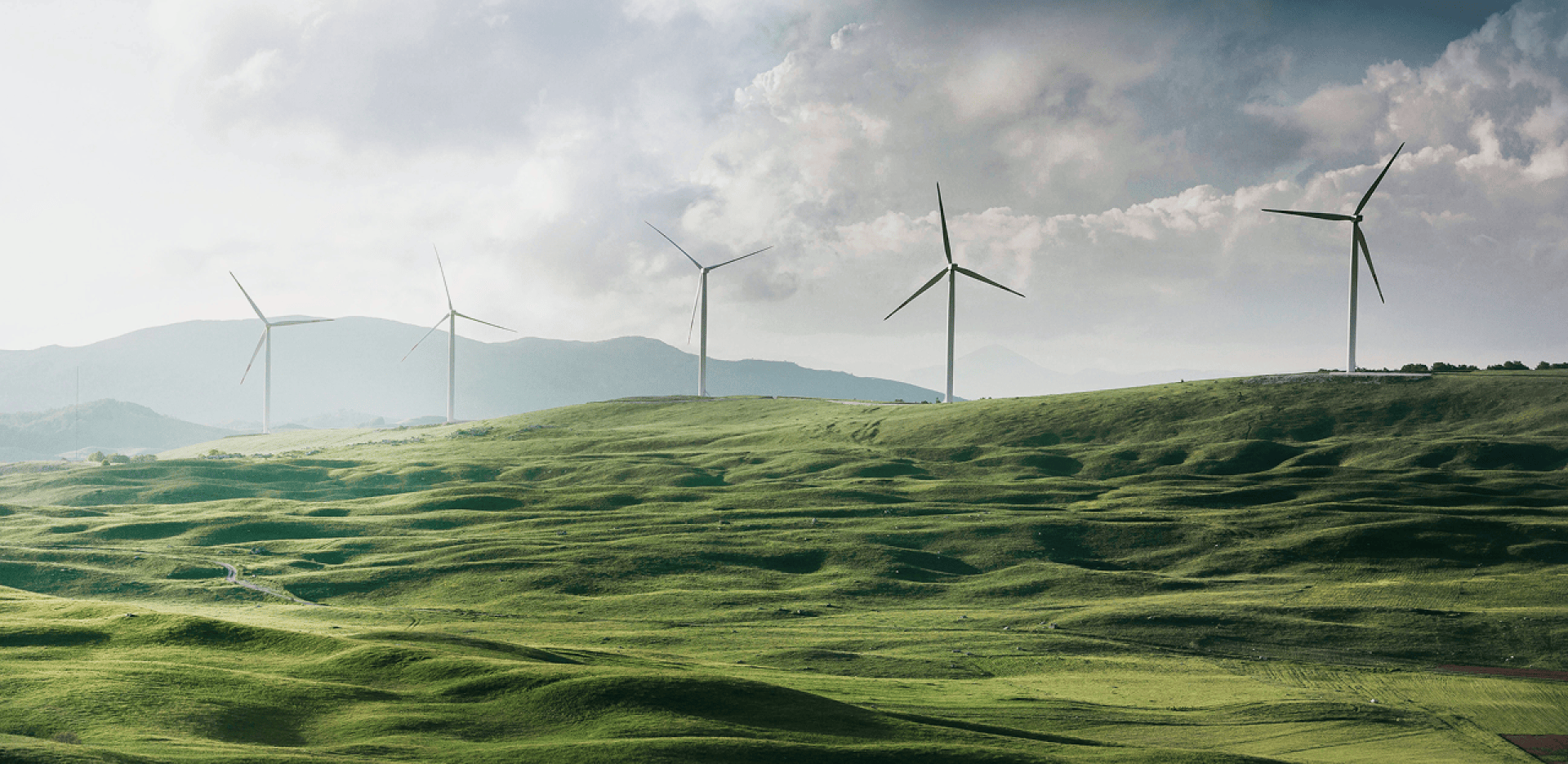 Wind turbines stand on rolling green hills under a cloudy sky, with distant mountains in the background, illustrating a landscape focused on renewable energy and sustainability.