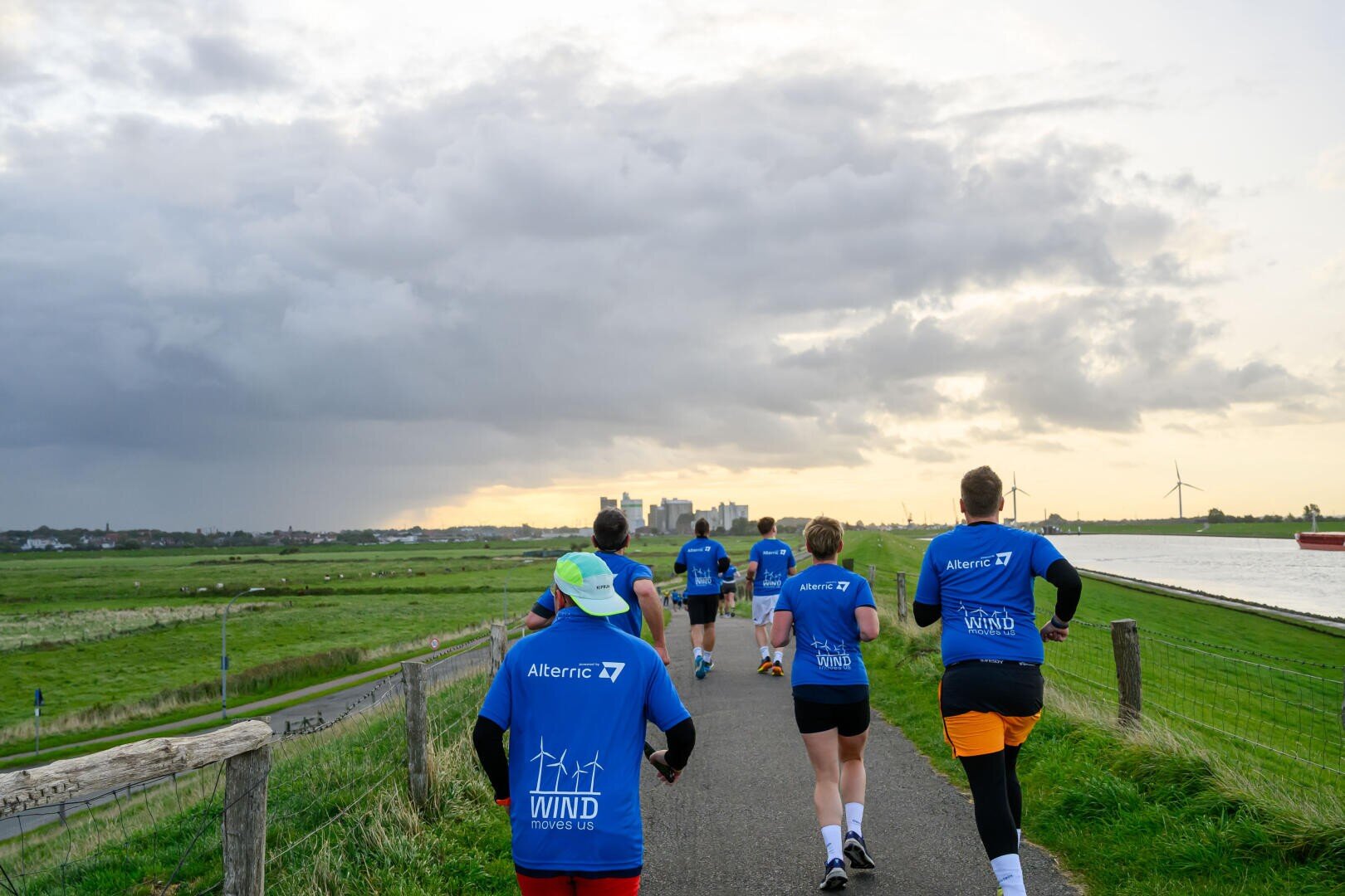 Eine Gruppe von Menschen in blauen Hemden joggt bei bewölktem Himmel und Sonnenuntergang einen gepflasterten Weg entlang eines Flusses mit grasbewachsenen Feldern und Windrädern auf beiden Seiten.