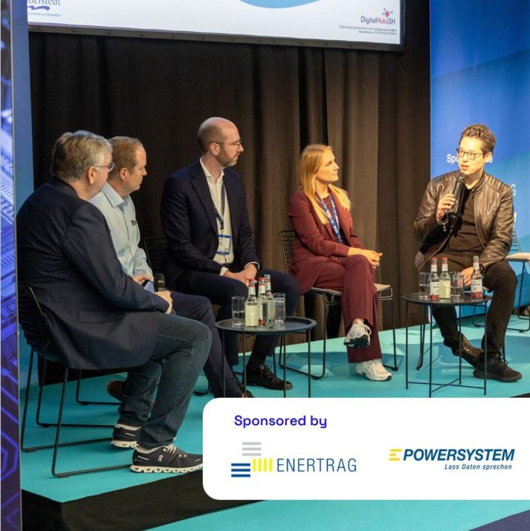 Five people sit on a panel discussion stage with microphones and water bottles. They are engaged in conversation. A sign in front of the stage reads “Sponsored by ENERTRAG and POWERSYSTEM.”.