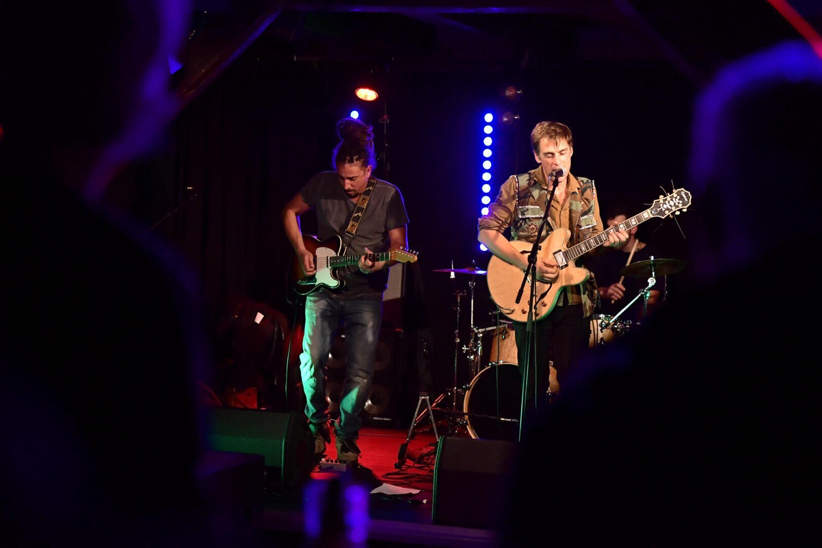 Two musicians perform on a dimly lit stage, one playing electric guitar and the other singing into a microphone while playing acoustic guitar; drums and stage lights can be seen in the background.
