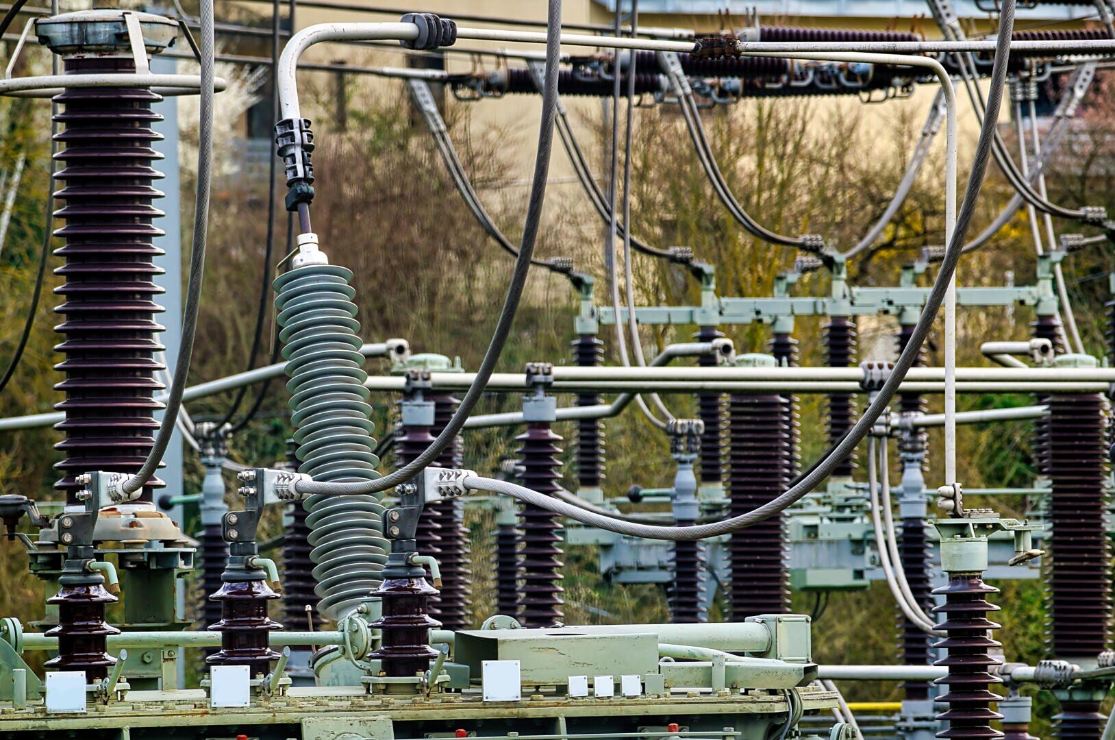 Close-up of an electrical installation in a substation with insulators, wires and metal components arranged in a complex outdoor network. Trees and a building can be seen in the blurred background.