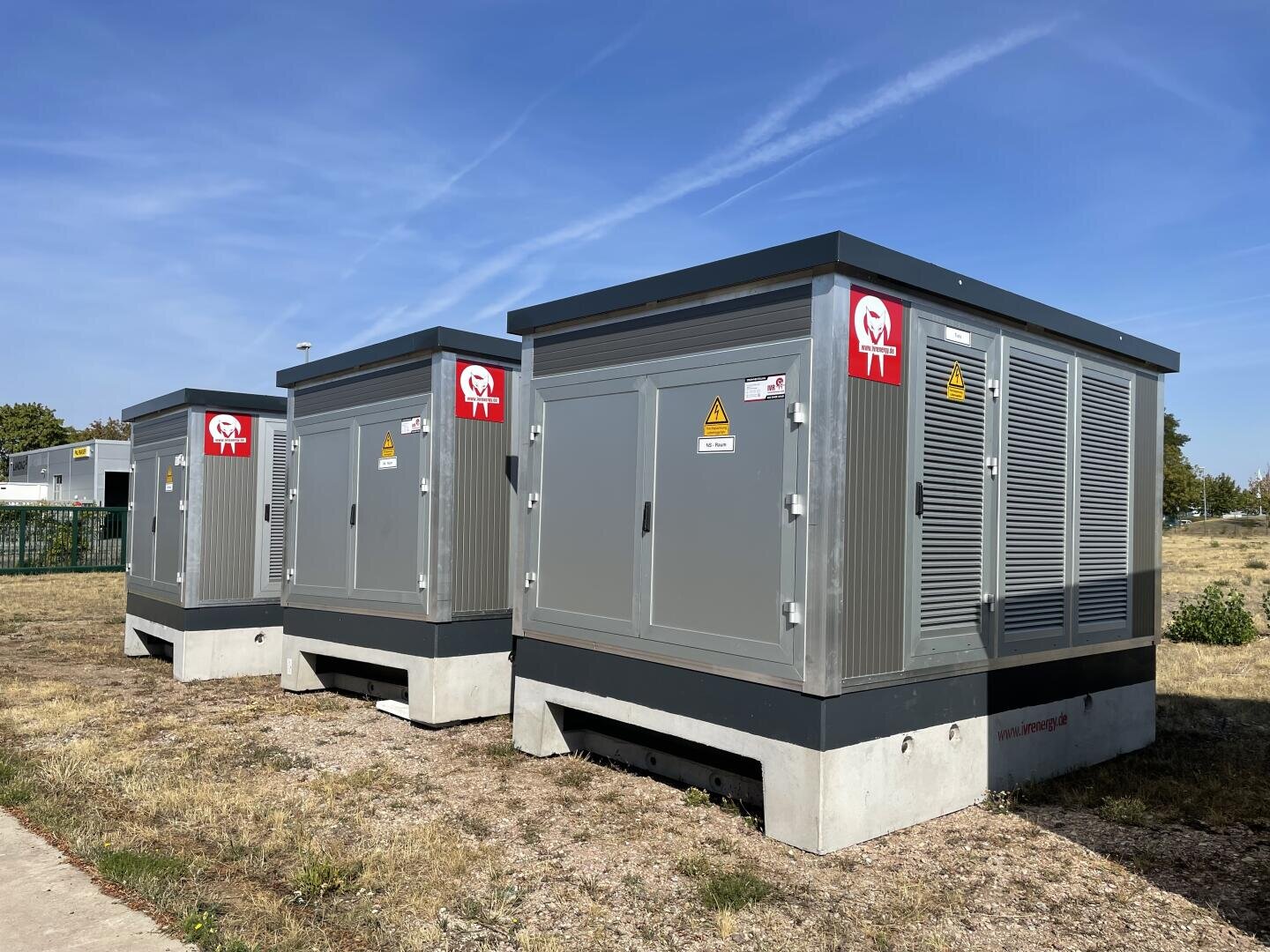 Drei große, graue Transformatorenkästen stehen im Freien auf Betonsockeln, die mit Warnschildern und rot-weißen Etiketten versehen sind, auf einer Wiese unter blauem Himmel.