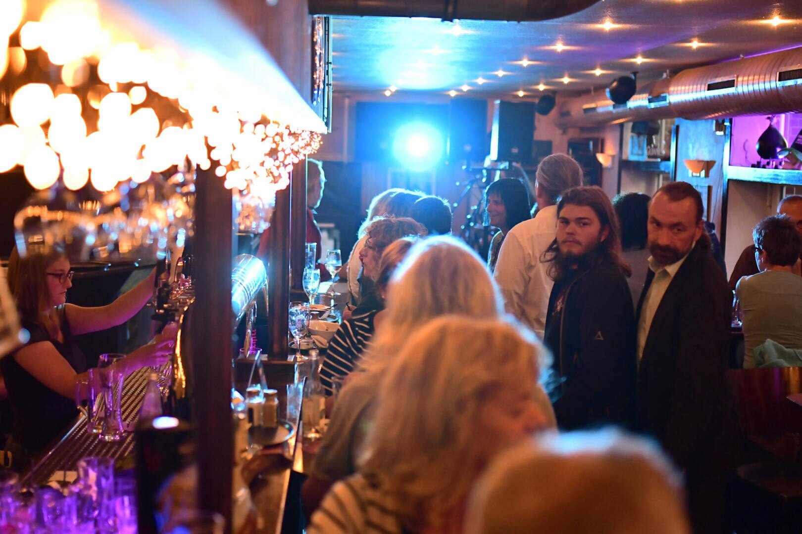 Eine gut besuchte Bar mit geselligen Gesprächen und an der Theke stehenden Gästen. Warmes Licht sorgt für eine gemütliche Atmosphäre, und ein hellblaues Licht strahlt aus dem hinteren Teil des Raumes und beleuchtet einige Gäste.