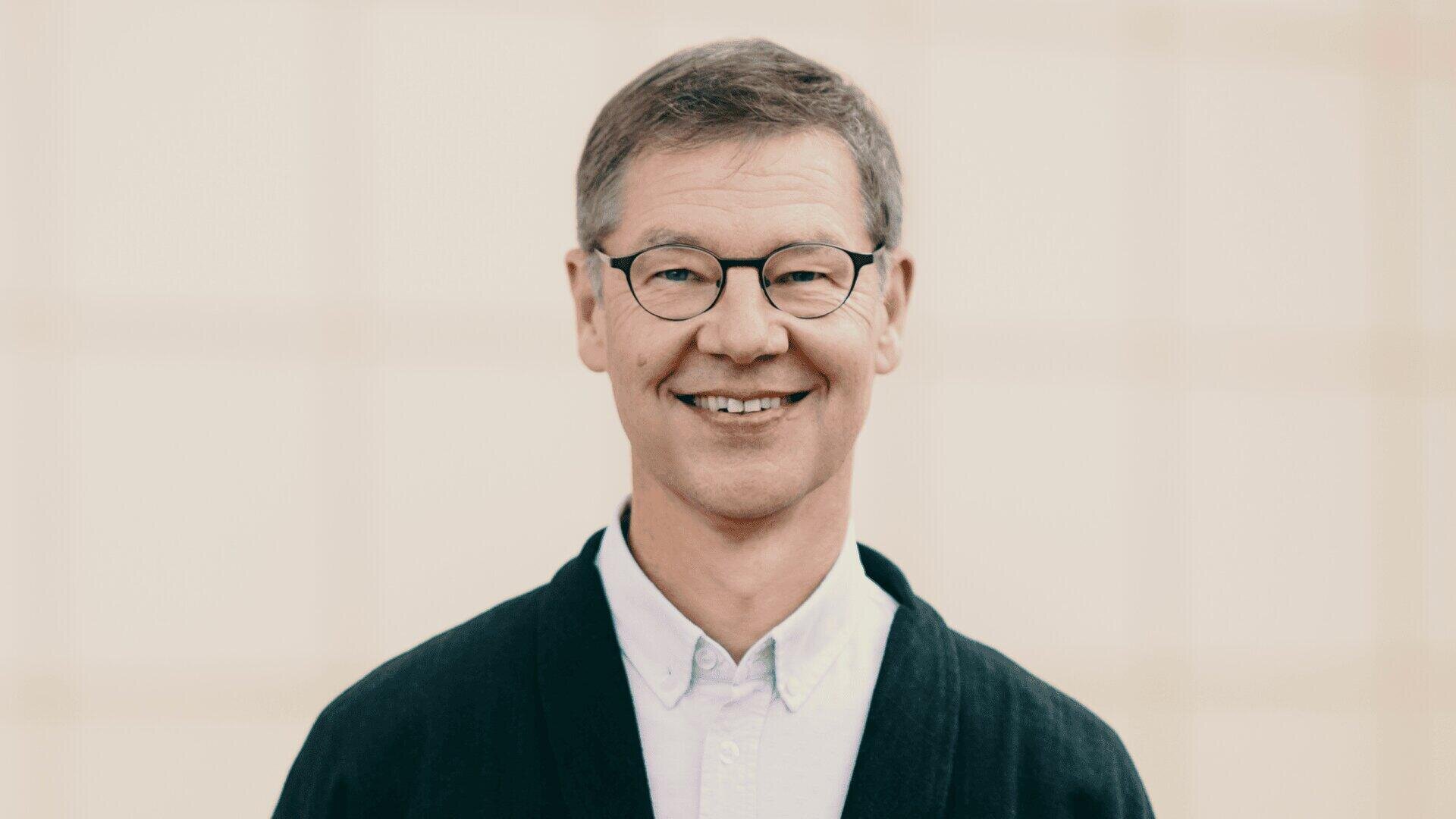 A middle-aged man with short brown hair and glasses smiles at the camera. He wears a white collared shirt and a dark sweater and stands in front of a bright, blurred background.