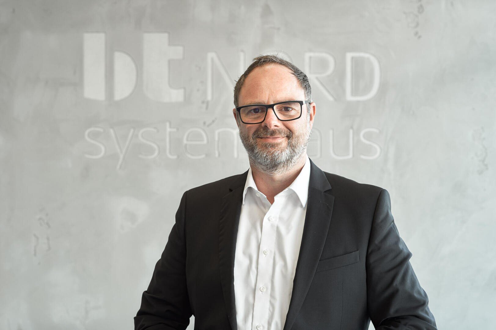 A man with glasses and a beard, wearing a black suit and white shirt, stands in front of a grey wall with the text bt NORD systemhaus partially visible in the background.