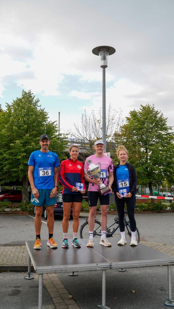 Vier Läufer stehen im Freien auf einem Podium und lächeln in die Kamera. Zwei Männer und zwei Frauen tragen Sportkleidung und Startnummern; ein Mann hält einen großen Pokal in der Hand. Im Hintergrund sind Bäume und ein Laternenpfahl zu sehen.