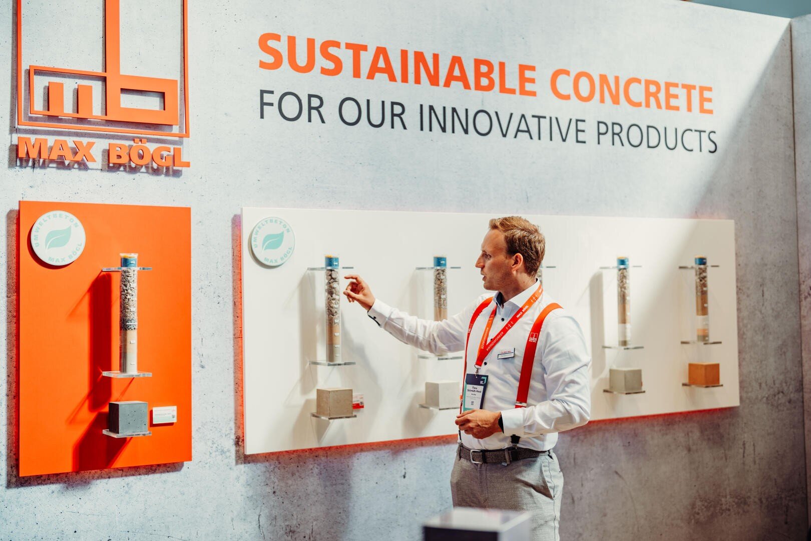 A man in a white shirt and red suspenders points to a wall with samples of sustainable concrete with the inscription SUSTAINABLE CONCRETE FOR OUR INNOVATIVE PRODUCTS at a Max Bögl trade fair stand.