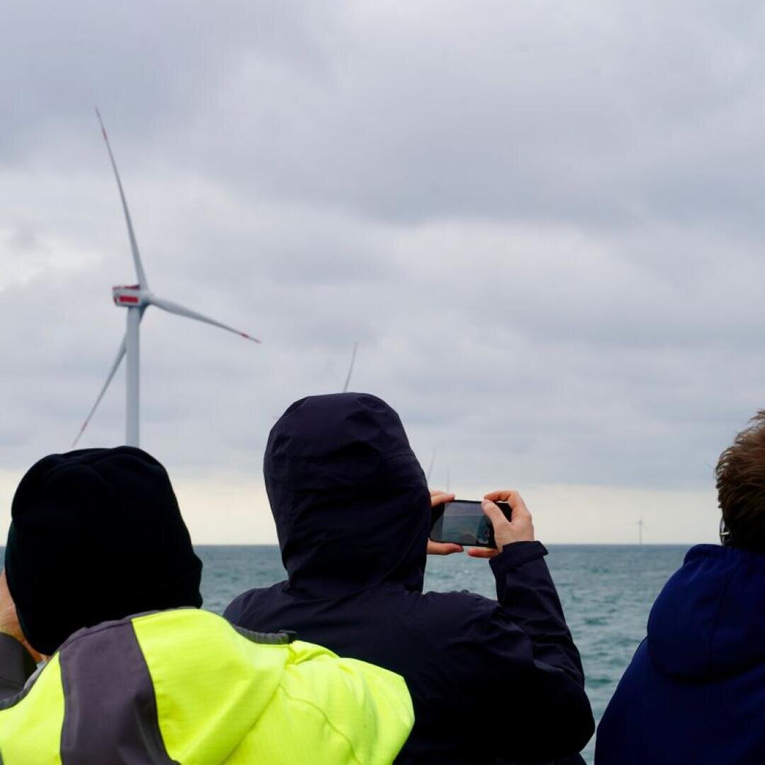 Drei Personen in Jacken blicken aufs Meer hinaus, eine davon macht mit ihrem Handy ein Foto. Unter einem bewölkten Himmel ragen große Windräder aus dem Wasser.