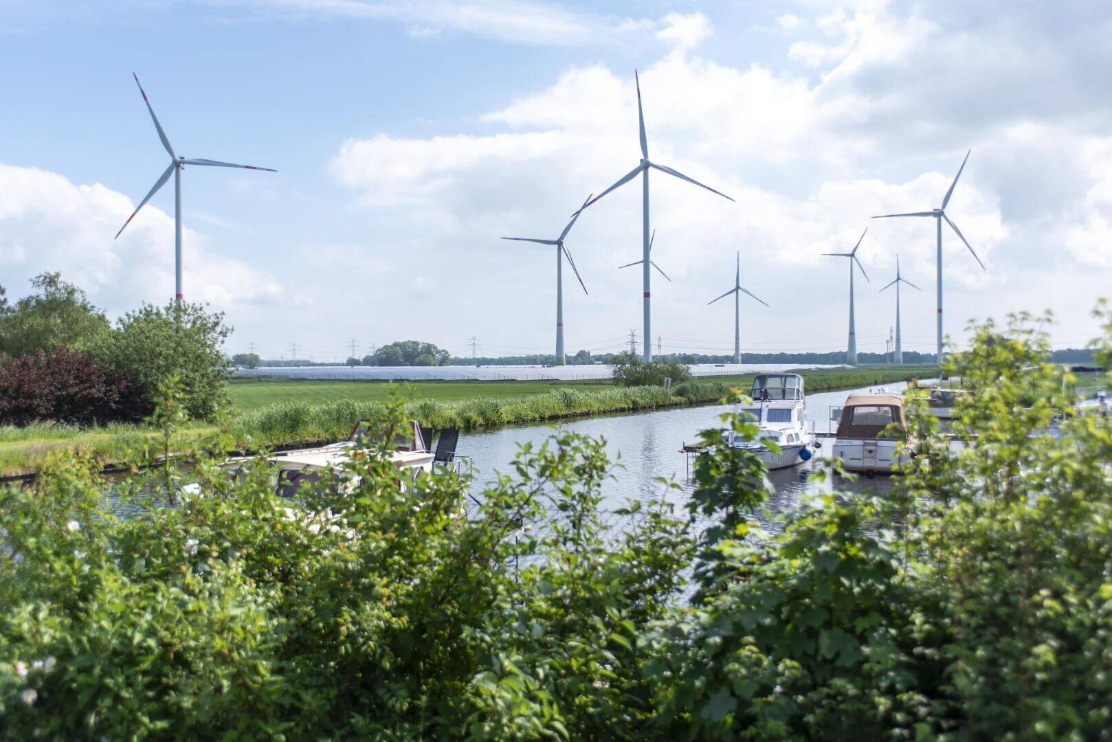 Windturbinen stehen auf einer grünen Wiese in der Nähe eines ruhigen Flusses, an dessen Ufer Boote angedockt sind, während üppige Büsche und Bäume den Vordergrund unter einem teilweise bewölkten Himmel einrahmen.