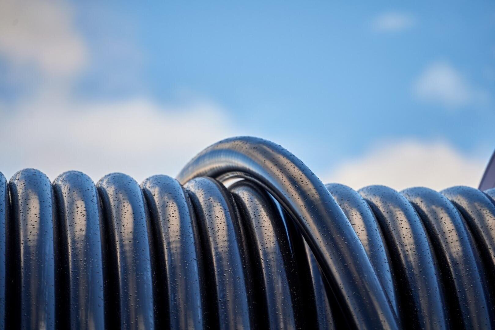 Nahaufnahme eines aufgerollten schwarzen Schlauchs mit Wassertropfen auf seiner Oberfläche vor einem blauen Himmel mit vereinzelten Wolken.