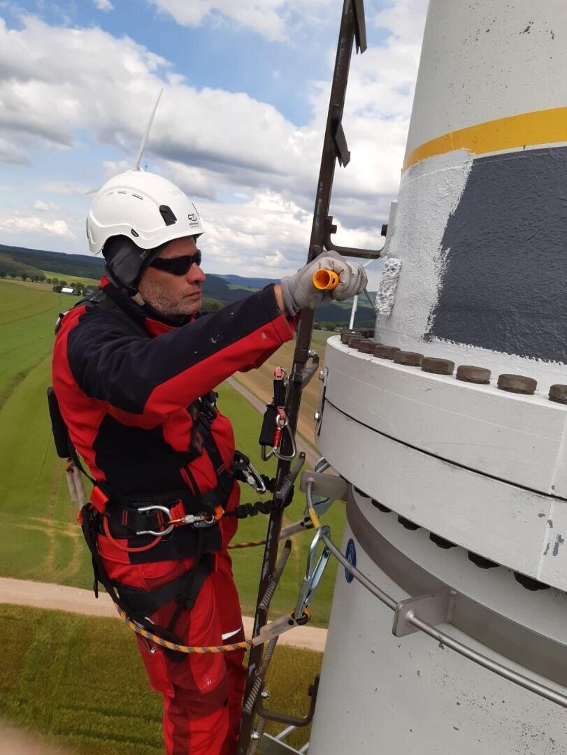 Ein Arbeiter, der Schutzkleidung und einen Helm trägt, streicht mit einer Farbrolle eine hohe Metallkonstruktion, möglicherweise einen Windradturm, während er mit Gurten über einer grünen Landschaft gesichert ist.