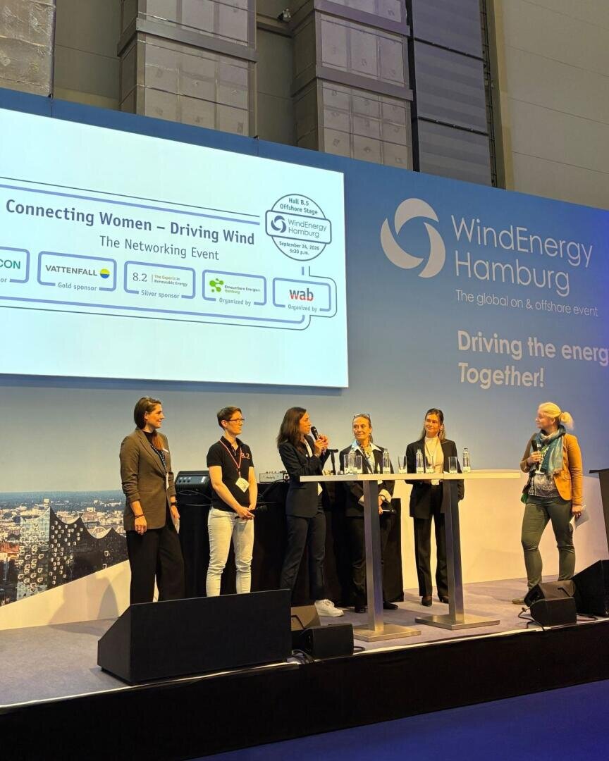 Sechs Frauen stehen auf der Bühne der Veranstaltung „WindEnergy Hamburg“ und halten eine Rede, unter einem Schild mit der Aufschrift „Connecting Women – Driving Wind“. Im Hintergrund sind Veranstaltungs- und Sponsorenlogos in den Farben Blau und Weiß zu sehen.