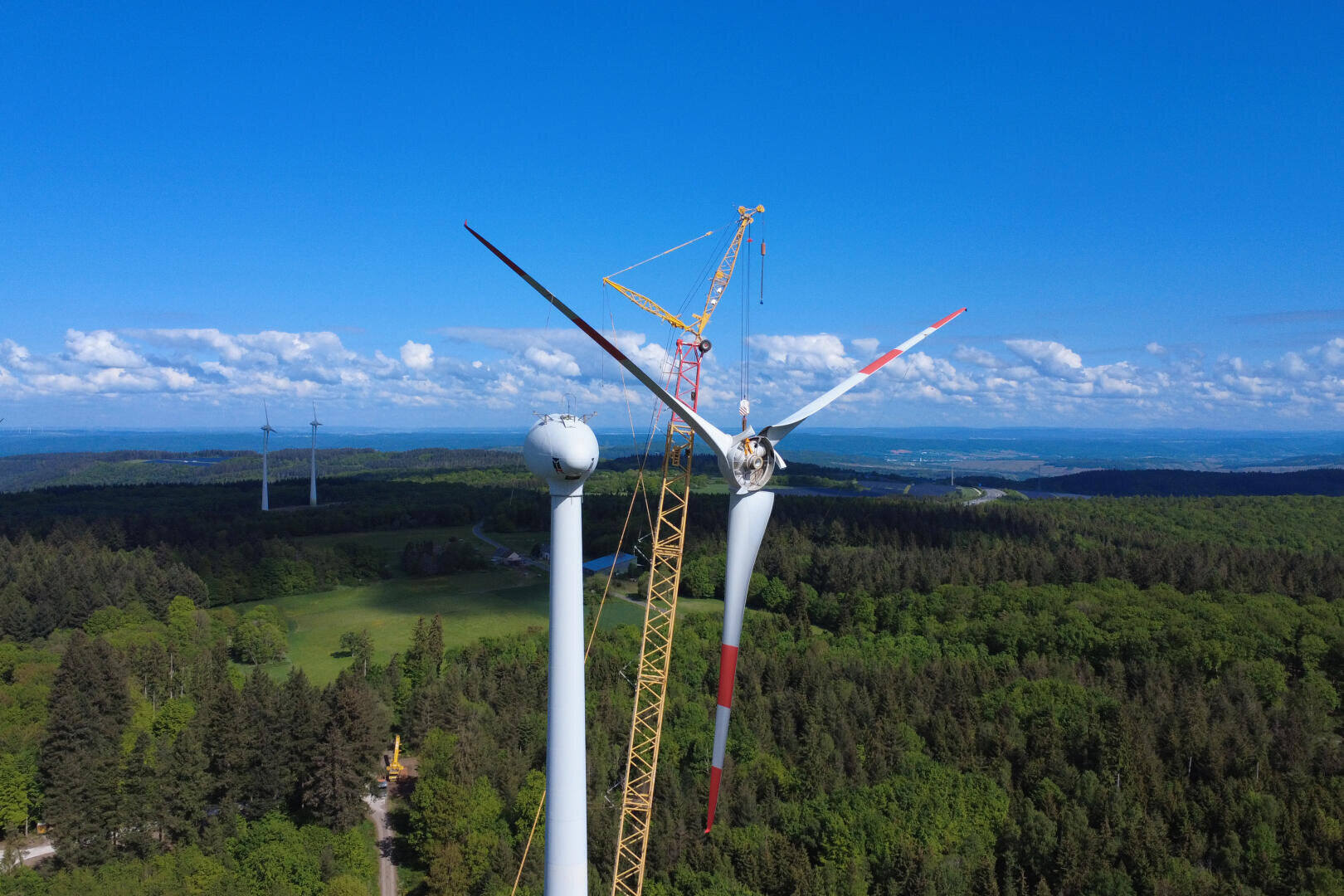 Ein Kran demontiert ein großes Rotorblatt an einer Windkraftanlage über einer bewaldeten Landschaft unter einem strahlend blauen Himmel mit vereinzelten Wolken. Im Hintergrund sind weitere Windkraftanlagen zu sehen.