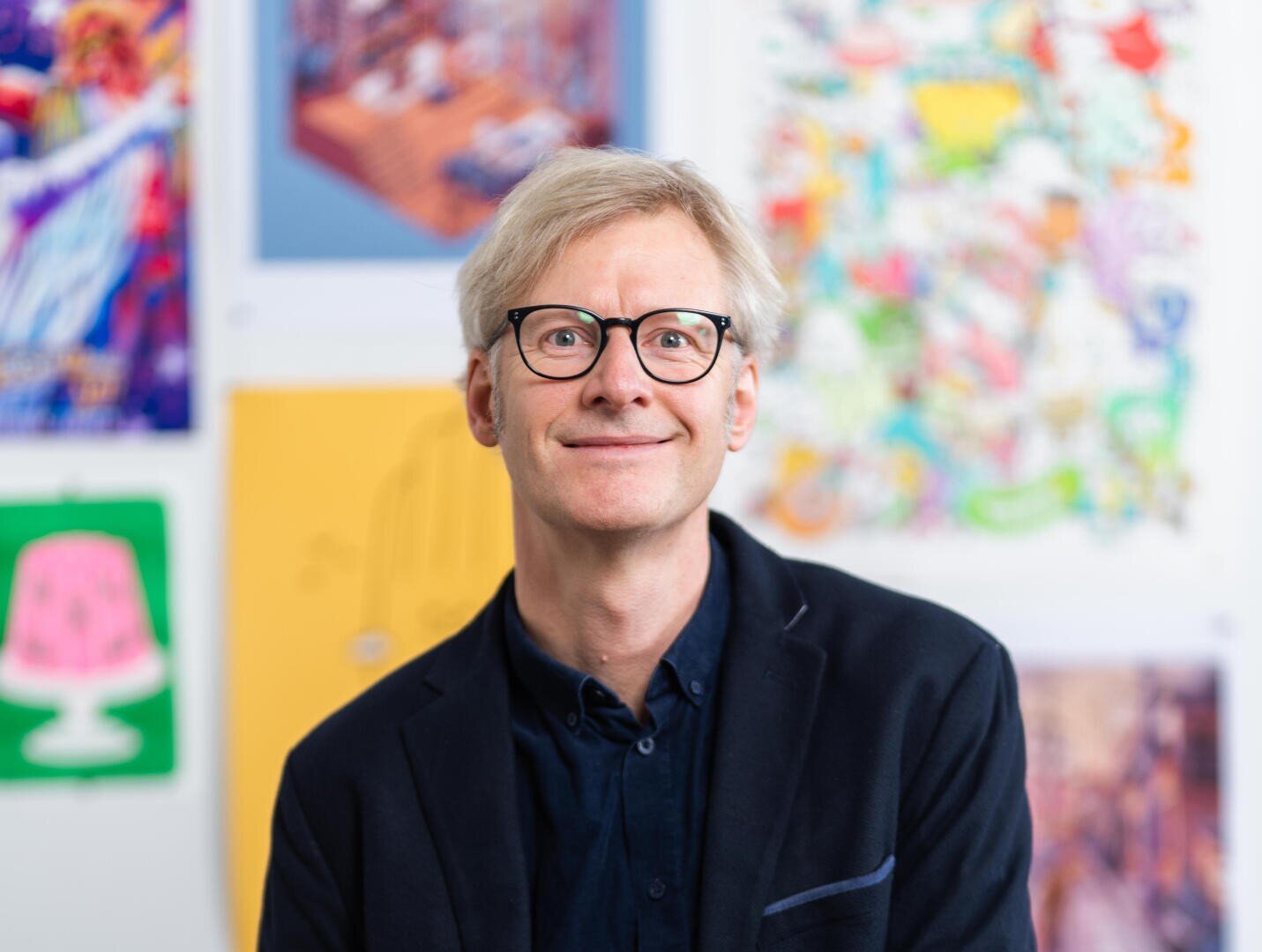 A smiling man with blond hair and glasses wearing a dark blazer, standing in front of a wall covered with colorful artworks and illustrations.