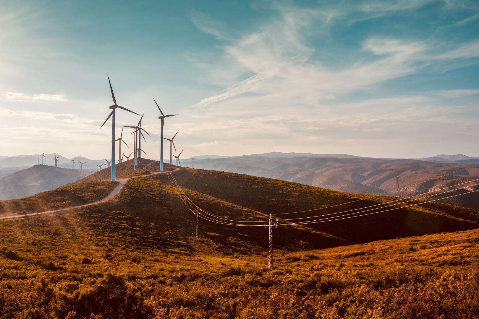 Windkraftanlagen stehen auf einem sonnenbeschienenen, mit Sträuchern bewachsenen Hügel, durch den Stromleitungen verlaufen. Im Hintergrund sind sanfte Berge bei teilweise bewölktem Himmel zu sehen.