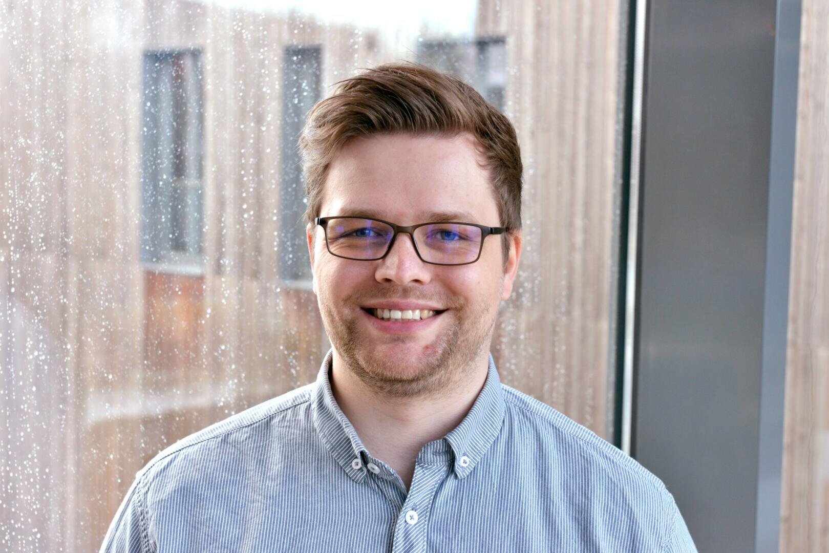 Ein lächelnder Mann mit Brille und kurzem, hellbraunem Haar steht in einem Innenraum an einem großen Fenster, auf das Regentropfen fallen, und trägt ein hellblau gestreiftes Button-up-Hemd. Draußen sind verschwommene Gebäude zu sehen.