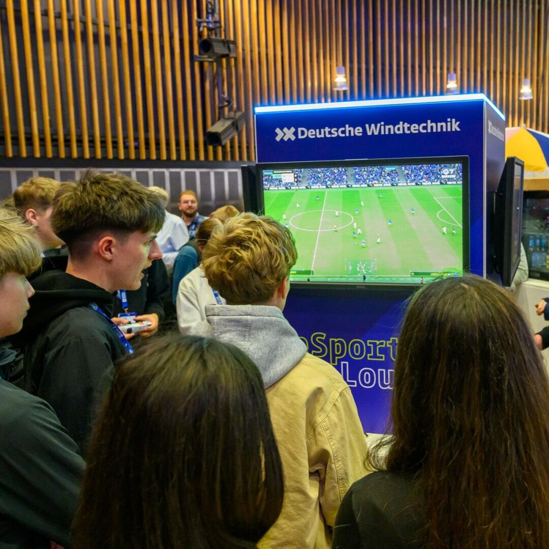 Eine Gruppe junger Leute schaut sich ein Fußball-Videospiel an, das auf einem großen Bildschirm an einem Indoor-Event-Stand mit der Aufschrift Deutsche Windtechnik gespielt wird.