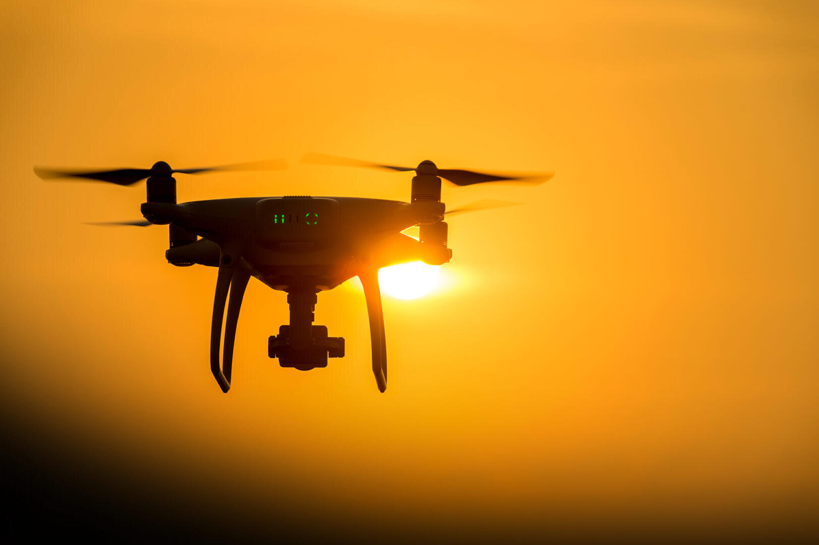 The silhouette of a drone in flight stands out against a bright orange evening sky, with the low sun partially visible behind it.