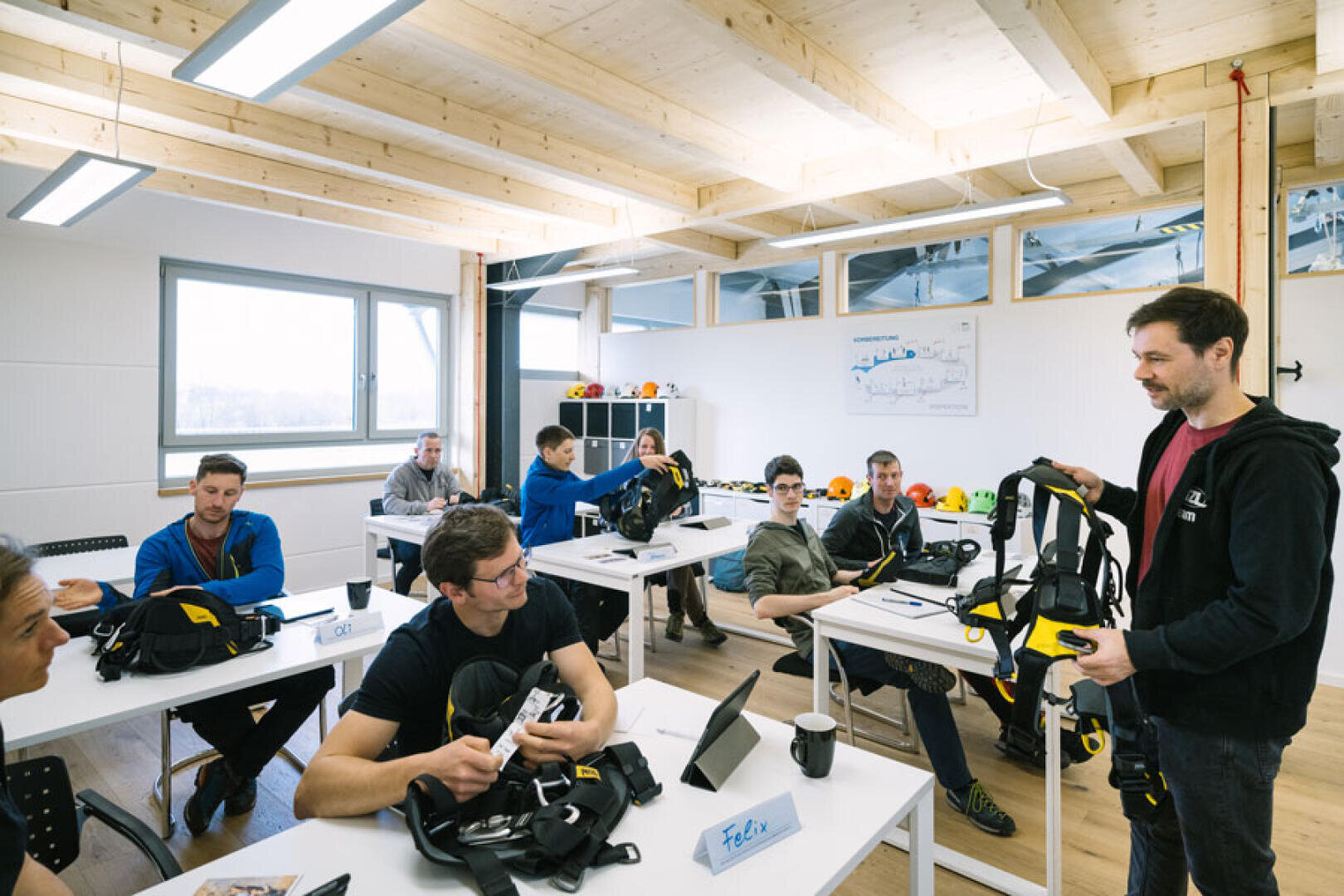 Ein männlicher Ausbilder demonstriert einer Gruppe von Schülern, die in einem hellen Klassenzimmer an Tischen sitzen, die Sicherheitsausrüstung. Die Schüler beobachten und bedienen ähnliche Geräte. Der Raum hat große Fenster und freiliegende Holzbalken.