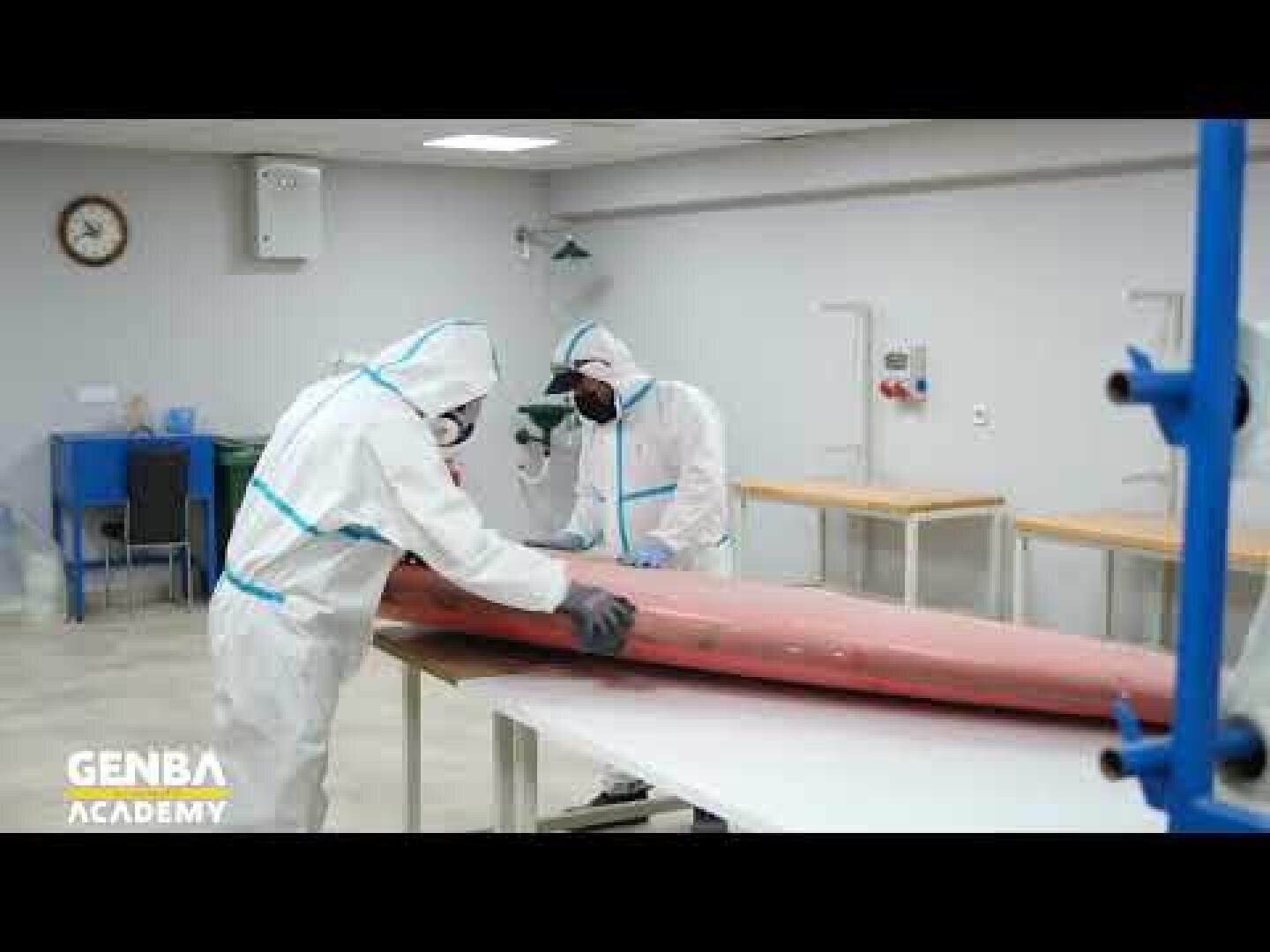 Two people in full protective suits and masks work on a large, elongated red object in a clean, laboratory-like room. The GENBA Academy logo is visible in the lower-left corner.