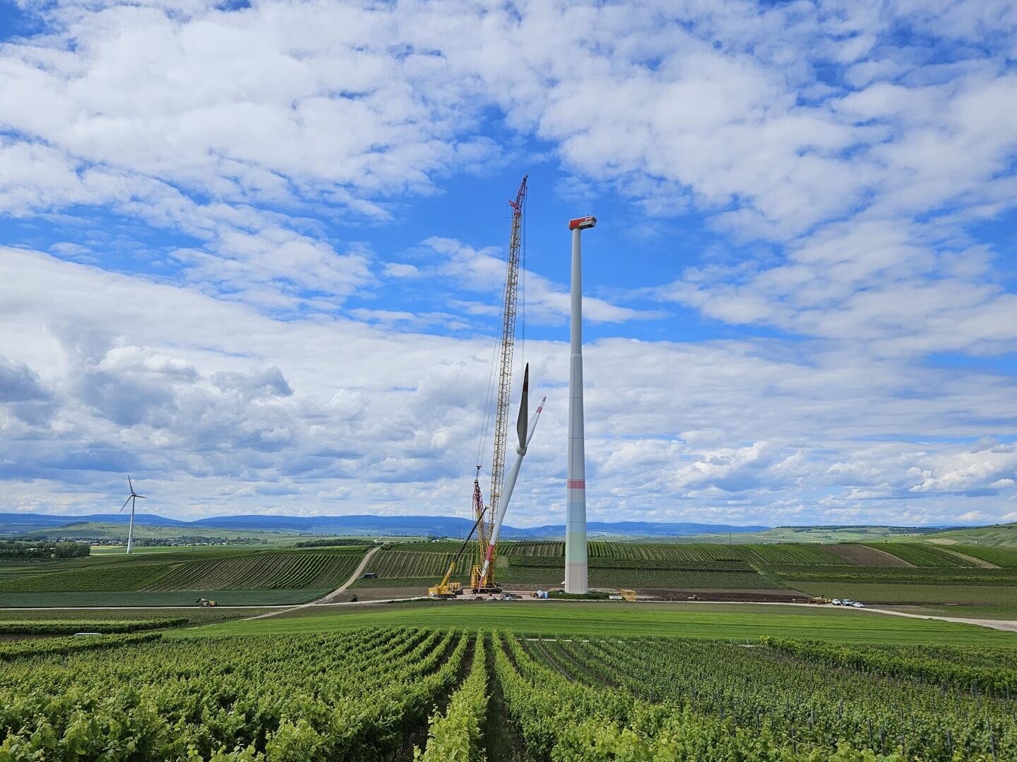 A wind turbine is being dismantled on a field, with cranes standing next to it. In the background, under a blue sky with scattered clouds, a turbine is not yet being dismantled.