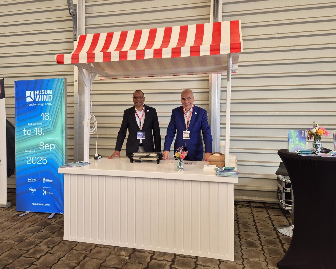Two men in suits stand smiling behind a white stand with a red and white striped canopy. A blue Husum Wind event banner and a black table with brochures stand next to them in an exhibition room.