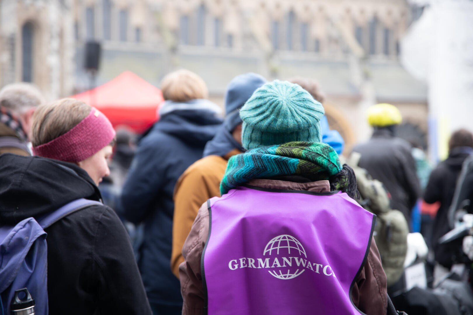 Eine Person, die eine blaue Strickmütze und eine lila Weste mit der Aufschrift Germanwatch trägt, steht inmitten einer Gruppe von Menschen im Freien, von denen einige Winterkleidung tragen. Der Hintergrund ist leicht unscharf.