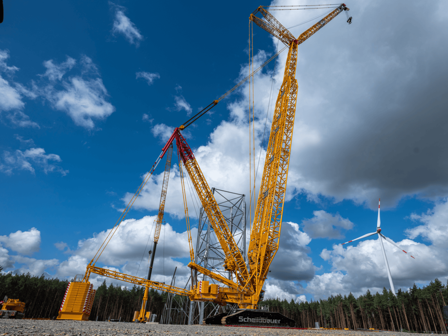 Ein großer gelber Baukran hebt Ausrüstung auf einer Baustelle unter blauem Himmel mit weißen Wolken. Im Hintergrund sind Bäume und eine Windkraftanlage zu sehen.