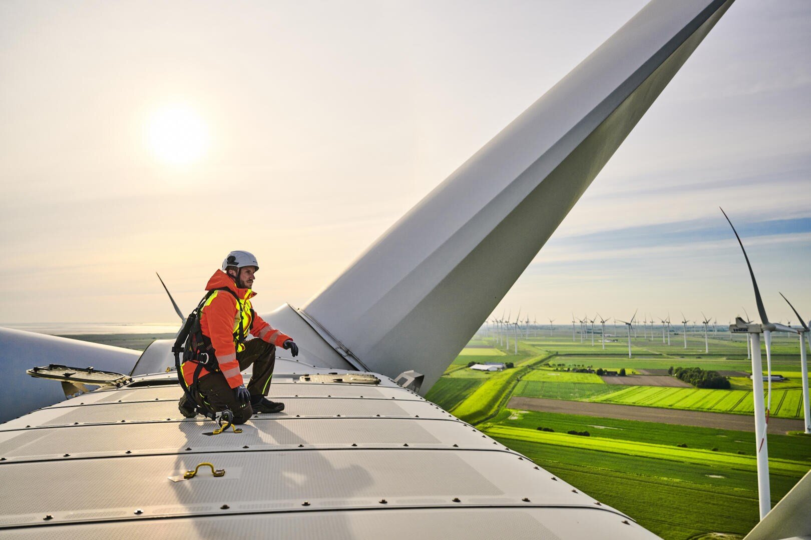A worker in protective clothing sits on a wind turbine overlooking green fields and other turbines under a bright sky while the sun is low on the horizon.