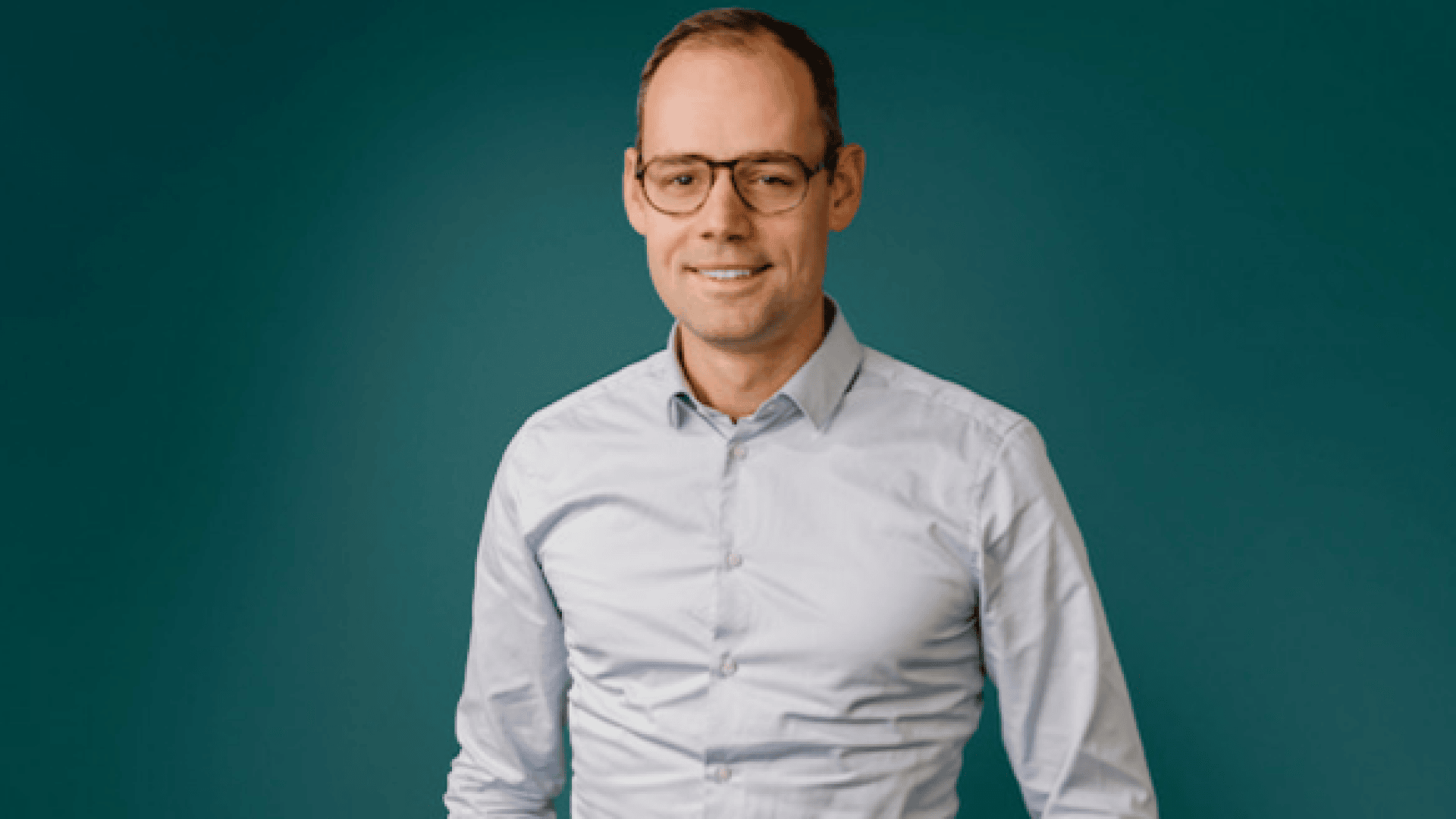 Ein Mann mit Brille und hellblauem Button-up-Hemd steht vor einem einfarbig türkisfarbenen Hintergrund und lächelt leicht in die Kamera.